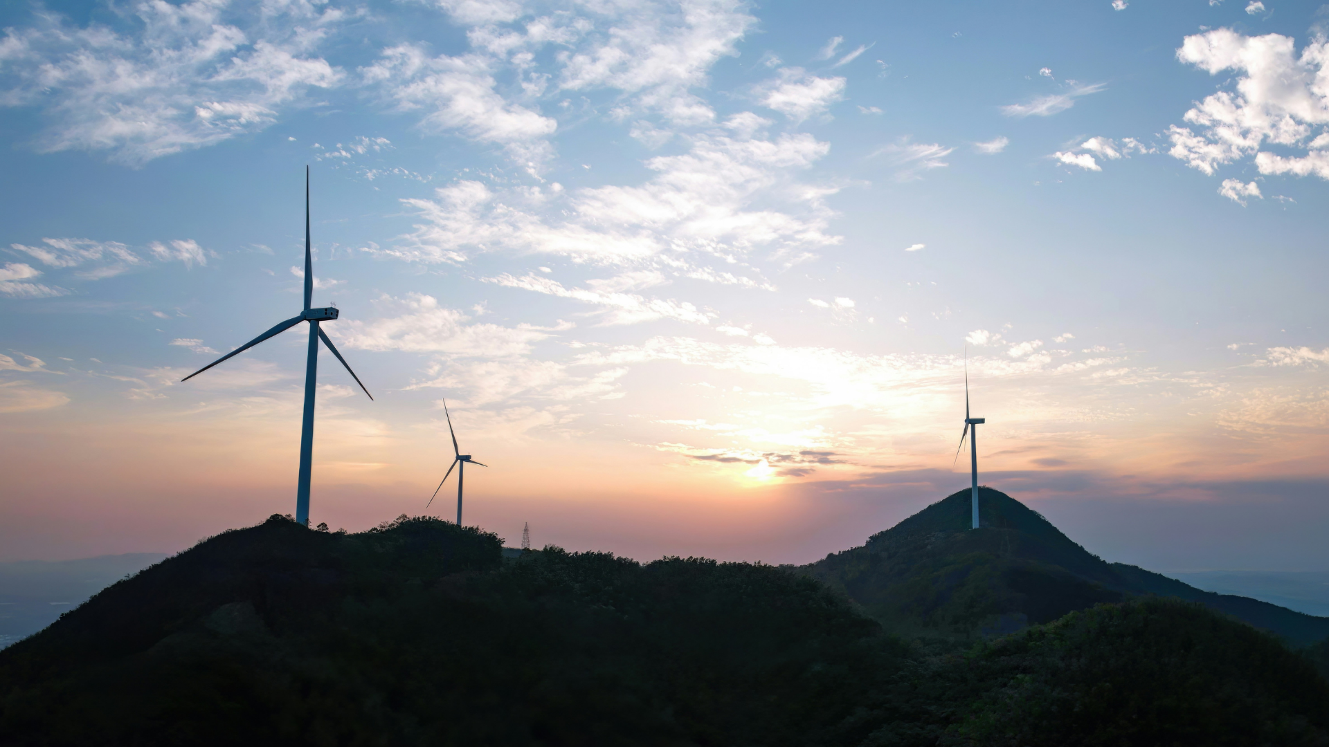 Three wind turbines on a hill, with a cloudy light blue sky. A sun appears on the horizon